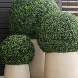 Drie grote Kunstplant Buxus romeinse stijl UV D65cm potten tonen netjes gesnoeide, bolvormige groene vormsnoei tegen een achtergrond van donkere houten latten op een houten vloer.
