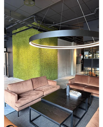 Een moderne zithoek met beige banken, zwarte salontafels, een grote ronde plafondlamp en op de achtergrond een groene muur, gecreëerd met Kunsthaag Nordic Mos 50x50 cm UV, die een luxe interieur en een eigentijds industrieel ontwerp uitstraalt.