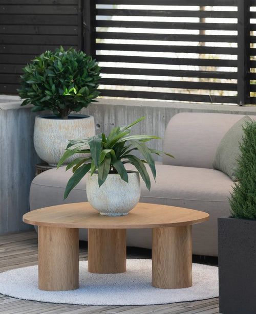 Op een klein vloerkleed naast een grijze bank op een terras staat een houten salontafel met daarop een Kunstplant Bird’s Deluxe van 60 cm, met op de achtergrond nog enkele potplanten.