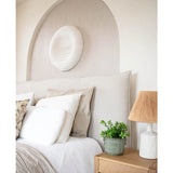 Een gezellige slaapkamer met een beige hoofdeinde, neutraal beddengoed en kussens, een houten nachtkastje met daarop de Kunstplant Eucalyptus 27 cm, een lamp met een getextureerde kap en ronde witte muurkunst boven het bed in een gewelfde nis.