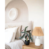 Een gezellige slaapkamer met neutrale tinten heeft een bed met wit linnen, een houten nachtkastje met de Kunst Begonia 48 cm in sierpot zwart en een geweven lamp, en ronde muurkunst met textuur boven het bed.