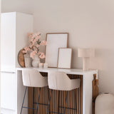Een minimalistische thuisbar met twee beige krukken met kussens, een witte toonbank, ingelijste kunst, een tafellamp, neutraal getinte decoratie en Kunst Kersenbloesem 100 cm licht roze in vazen tegen een lichtgekleurde muur.
