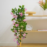 Een houten plank met Kunst hangplant Petunia 70 cm paars, een witte klok, een houten standaard, een boek en een vaas met roze bloemen tegen een witte muur.