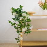 De Kunst hangplant Petunia 70 cm wit met witte bloemen valt van een houten schap in een lichte, moderne kamer met lichte houten vloeren. Op de bovenste plank staat een vaas met lange roze bloemen.