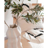 Een wit gebeeldhouwde vaas houdt Kunst olijfblad 90 cm UV met groene bladeren op een lichte houten tafel. Daarachter versterken een beige stoel en twee crèmekleurige kussens met opvallende zwarte strepen de mediterrane sfeer.