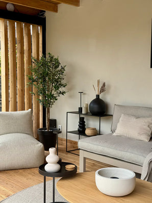 Een moderne woonkamer met lichtbeige muren, grijze bank, zachte fauteuil, zwarte bijzettafeltjes met vazen, houten vloer en een Kunst olijfboom 160 cm UV potplant. Verticale houten latten zorgen voor natuurlijk licht vanaf de zijkant.