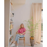 Een vrouw in een roze trui en een spijkerbroek zit op een houten stoel naast een Kunst varen-plant van 120 cm, met boekenkasten en een raam met beige gordijnen in een lichte, moderne kamer.