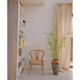 Naast een 120 cm hoge Kunst varen in een pot staat een houten stoel, vlakbij crèmekleurige gordijnen en een muur met planken vol boeken en decoratie. Zonlicht verlicht de minimalistische kamer, waar stijlvolle kamerplanten zoals de Kunst varen een vleugje door de natuur geïnspireerde rust toevoegen.