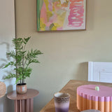 In een kamer met een pastelthema staan een Kunst-varenplant van 85 cm op een roze bijzettafeltje, een paars-gouden kopje, een roze schaal met geschulpte rand, een open notitieboekje en aan de muur een abstract schilderij met groene, roze en gele tinten.