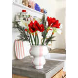 De Kunstbloem Amaryllis Real Touch rood 36 cm staat in een wit met roze gestreepte vaas van keramiek op een betonnen aanrecht in een lichte keuken met planken met kleurrijk servies op de achtergrond.
