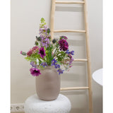 Een Kunstbloem Grasklokje van 62 cm paars staat in een matglazen vaas op een gestructureerde witte sokkel. Daarachter leunt een lichte houten ladder tegen een beige muur, met stopcontacten zichtbaar bij de vloer.