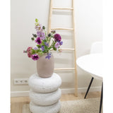 Een Kunstbloem Grasklokje van 62 cm paars staat in een vaas op drie ronde, witte steenachtige kussens. Daarachter staat een lichthouten ladder tegen een witte muur, naast een witte tafel en stoel op een beige vloerkleed.