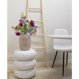 Een Kunstbloem Grasklokje 62 cm paars staat in een beige vaas op gestapelde witte terrazzo krukjes. Daarachter staat een lichthouten ladder, bij een witte stoel en een ronde tafel, allemaal geplaatst op een beige vloerkleed met textuur in een minimalistische kamer.