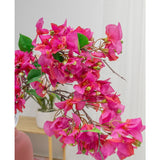 Een close-up van de Kunstbloem Bougainvillea 90 cm paars toont zijn levendige paarse bloemen en groene bladeren binnenshuis, met een potplant op een houten plank en een witte muur zacht vervaagd op de achtergrond.