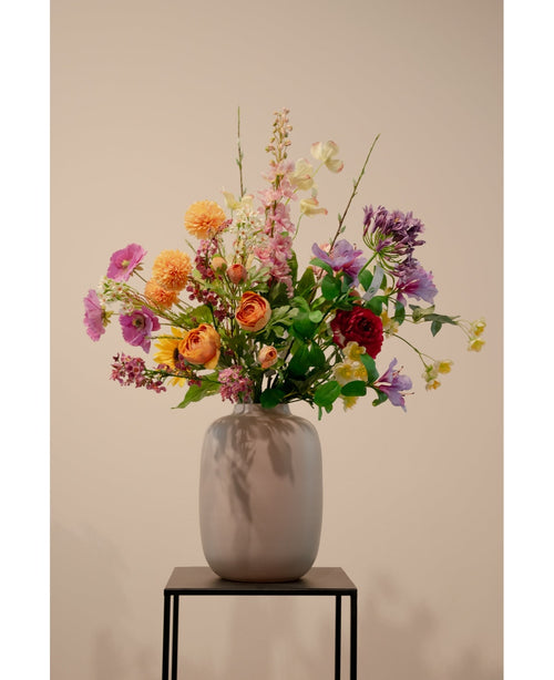De Kunstkornoelje Real Touch Lichtgeel 74cm, een levensechte lichtgele kunstbloem, staat in een keramieken vaas met groen gebladerte op een zwart metalen standaard tegen een effen beige achtergrond.