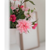Een close-up van de Kunstbloem Dahlia 68 cm roze in een witte vaas, met een vaag fotolijstje op de achtergrond op een wit vlak.