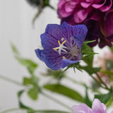 Een close-up van het Kunstbloem Grasklokje 62 cm paars, een delicate paarse kunstbloem met diepe nerven en gele meeldraden, tussen groene bladeren en zacht vage roze en paarse bloemen op de achtergrond.