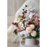 De Kunstbloem Jilly 70 cm roze kunstbloem staat in een beige vaas op een houten vloer naast een zacht ivoorkleurig schapenvachtkleed.