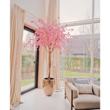 De Kunstboom Kersenbloesem roze 240 cm staat in een decoratieve pot bij een groot raam in een lichte, moderne woonkamer met crèmekleurige gordijnen, een beige fauteuil en uitzicht op de tuin buiten.