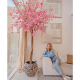 Een vrouw in casual kleding ontspant op een beige bank naast een 240 cm Kunstboom Kersenbloesem roze met roze bloemen, geplaatst in een moderne woonkamer met abstracte muurkunst en grote ramen.
