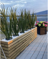 Een modern dakterras heeft een lange houten plantenbak met Kunstgras Groen UV 90cm en een zwarte vierkante plantenbak met kleurrijke hortensia's, allemaal op een lichtgrijs terras en omsloten door een glazen reling.