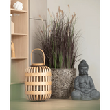 Een houten lantaarn, een hoge stenen pot met Kunstgras-uiengras-bonsai van 100 cm (brandvertragend) en een grijs Boeddhabeeld staan opgesteld op een lichtgekleurd oppervlak, vlakbij een houten plank en een bleke muur.