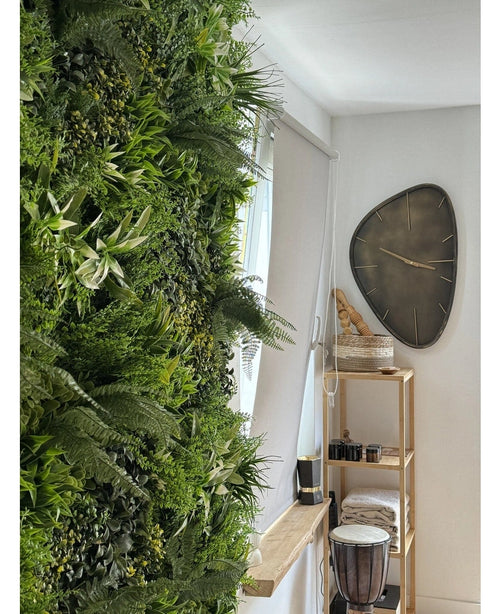 Een weelderige Kunsthaag Freshness 100x100 cm UV verticale tuin bedekt één muur van een lichte kamer. Vlakbij hangt een moderne driehoekige wandklok boven een houten plank met handdoeken, een djembé trommel, manden en kleine decoratieve stukken.