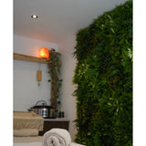 Een rustige spa-ruimte met een massagetafel, rustgevende verlichting, een Himalayazoutlamp op een plank en een weelderige Kunsthaag Freshness 100x100 cm UV groene plantenwand voor extra sereniteit.