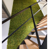 Een moderne trap met houten treden en zwarte leuningen staat naast een groene muur van Kunsthaag Nordic Mos 50x50 cm UV en voegt een natuurlijk, gestructureerd accent toe aan het luxe interieur.