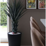 De Kunstplant Agave Pandora UV 120 cm, een hoge stekelige groene plant in een zwarte geribbelde pot met witte stenen, staat bij het raam. Daarachter hangen drie ingelijste fruitprints op een wandplank boven beige stoelen en een witte tafel.