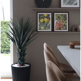 Een moderne eethoek met beige stoelen, een witte tafel, een wandklok en een plank met ingelijste foto's van citroen en bladeren. De grote Kunstplant Agave Pandora UV 120 cm voegt groen toe. Natuurlijk licht valt binnen vanaf de linkerkant.