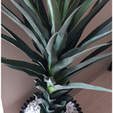 Een close-up van de Kunstplant Agave Pandora UV 120 cm, met groene stekelige bladeren in een zwarte pot met witte kiezels, uitgestald op een licht oppervlak tegen een neutrale muur.