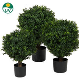 De Kunst Buxus op stam UV 40 cm heeft drie kunst vormbomen met dicht groen gebladerte in zwarte potten, die bij elkaar staan. Een klein UV-symbool met een zonnepictogram in de linkerbovenhoek geeft aan dat ze UV-bestendig zijn.