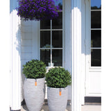 Twee Kunst Buxus op stam UV 40 cm ronde, gestructureerde grijze plantenbakken met groene struiken staan op een veranda voor een raam en deur. Daarboven siert een hanging basket met kleine paarse bloemen het witte huis met zuilen.