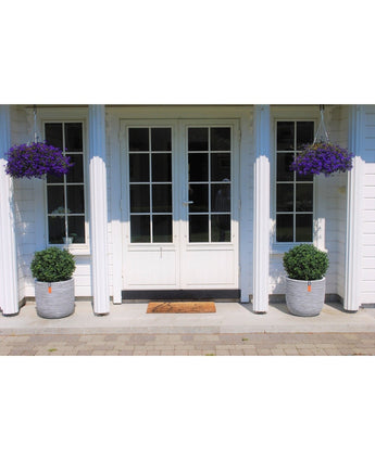 Witte dubbele deuren met glazen ruiten en witte zuilen worden geflankeerd door twee Kunstplant Buxus op stam romeinse stijl 65cm in potten. Daarboven hangen manden met paarse bloemen en voor de ingang ligt een welkomstmat.