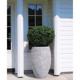 De Kunstplant Buxus op stam romeinse stijl UV 95cm, een hoge en realistische groene struik in een gestructureerde ronde grijze plantenbak, is geplaatst voor een wit houten huis met een raam en een glazen deur.