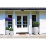 Voorportaal van een wit huis met dubbele deuren, zuilen en een hangende paarse bloemenmand. Drie Kunstplant Buxus op stam romeinse stijl UV 95cm plantenbakken met groene struiken staan aan weerszijden van de ingang.