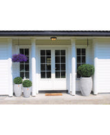 Een witte huisentree met dubbele glazen deuren is voorzien van twee Kunstplant Buxus op stam romeinse stijl 95cm plantenbakken, een welkomstmat, twee kleinere bijpassende plantenbakken, een hangende mand met paarse bloemen en vier witte zuilen die de deuropening omlijsten.