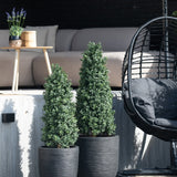 Twee Kunstplant Buxus Pyramide UV 90 cm vormbomen staan op een patio bij een buitenbank en een ronde rieten hangstoel met een zwart kussen, terwijl een klein tafeltje met een plant op de achtergrond staat.