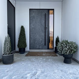 Een moderne donkere voordeur wordt omlijst door witte muren en geflankeerd door Kunstplant Buxus Pyramide UV 90 cm, die UV-bescherming biedt. Lichte sneeuw bedekt de grond, terwijl deze stijlvolle tuinplanten een opgeruimde, minimalistische ingang creëren.