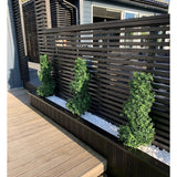 Drie Kunst Buxus Spiraal Romeinse stijl UV 90cm vormsnoeistruiken staan in een smal bed van witte kiezels, tegen een modern zwart privacyhek met latten naast een houten terras en een eigentijds huis.