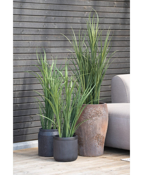 Een Kunstgras Groen UV 90cm kunstplant in een zwarte pot staat op een houten vloer naast een grijze buitenbank, met een donkere houten lattenwand op de achtergrond.
