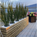 Een houten plantenbak met Kunstgras Groen UV 90cm en witte stenen staat op een houten vlonder naast een zwarte plantenbak met kleurrijke hortensia's, met glazen balustrades en uitzicht op de bergen op de achtergrond.