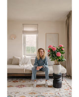 Een vrouw in spijkerpak zit op een beige bank in een lichte woonkamer. Op de voorgrond staat de Kunstplant Hortensia UV 85 cm roze, een grote roze kunsthortensia in pot op een zwarte standaard. Zonlicht stroomt door het raam achter haar.
