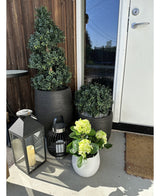 Een Kunstplant Hortensia Groen UV 35 cm in een witte pot met geelgroene bloemen staat op de veranda bij een bruine deur, naast twee planten in een zwarte pot en twee decoratieve lantaarns met kaarsen.