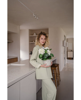 Een vrouw met krullend blond haar, gekleed in een lichtgroene outfit, staat in een moderne keuken met een Kunstplant Hortensia Wit UV 35 cm in haar handen – met crèmewitte bloemen – en glimlacht naar de camera.