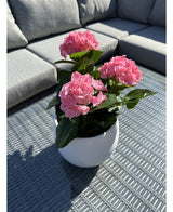 De Kunstplant Hortensia Roze UV 35 cm, een witte pot met levendige roze hortensiabloemen, staat op een geweven grijze buitentafel met een grijze sofa met kussens op de achtergrond, verlicht door zonlicht en zachte schaduwen.