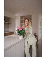 Een vrouw in een lichtgroen pak staat in een moderne keuken en glimlacht terwijl ze de Kunstplant Hortensia Roze UV 35 cm op het aanrecht vasthoudt.