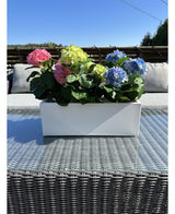 De Kunstplant Hortensia Roze UV 35 cm, een rechthoekige witte plantenbak met roze hortensia's, staat buiten op een rieten tafel met glazen blad, grijze kussens en een blauwe lucht op de achtergrond.
