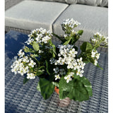 De Kunstbloem Kalanchoë plant UV 31 cm groot, met trosjes kleine witte bloemen en groene bladeren, staat op een rieten buitentafel met een grijze sofa met kussens op de achtergrond.